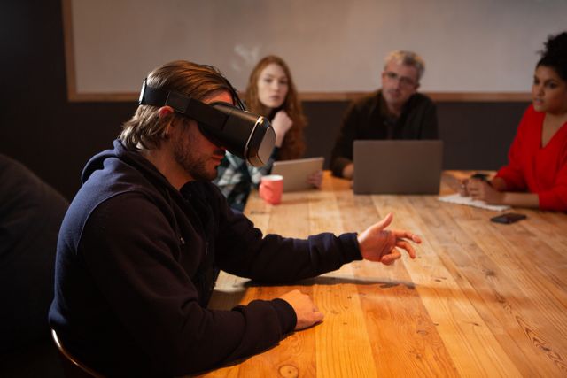 Side view of a Caucasian man working in a creative office, sitting by a table in a conference room, wearing a virtual reality headset, with his coworkers in the background
