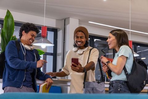 Diverse students laughing while sharing smartphone in bright modern campus lounge