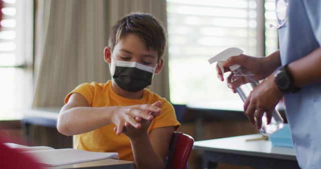 Boy applies hand sanitizer while wearing a face mask in a classroom. Ideal for topics on child safety, hygiene practices during COVID-19, school health protocols, and back-to-school season.