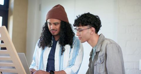 Two young men collaborating over laptop on angled wooden stand in bright creative studio