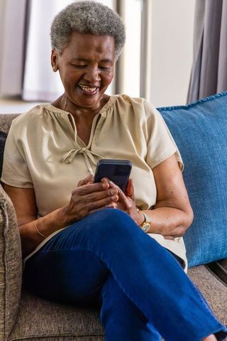 Senior african woman smiling while using smartphone on sofa, relaxed bright home moment
