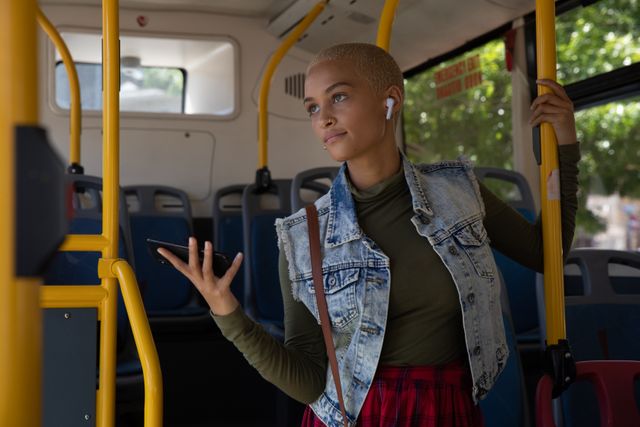 Biracial alternative woman with short blonde hair and denim jacket out and about in the city on a sunny day, with wireless earphones commuting on bus. Urban independent woman on the go.