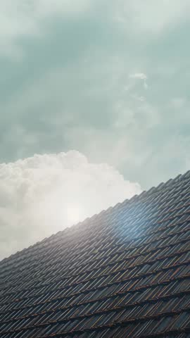 Tilting camera capturing sunlit rooftop tiles with cumulus cloud and lens flare in vertical video