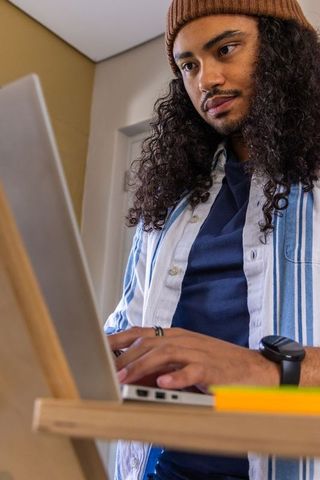 Young man typing on laptop at home workspace wearing brown beanie and striped shirt