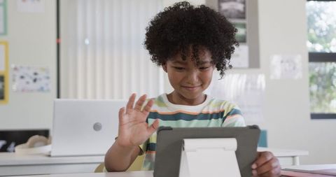 Young student waving during virtual lesson while focusing on tablet at classroom desk