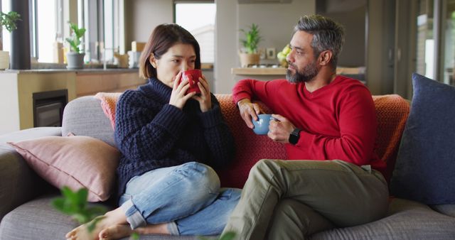 A couple sits on a sofa in a cozy living room, engaged in conversation while sipping coffee. The living space appears warm and inviting, with soft lighting and comfortable furnishings, including cushions and a knit blanket. Ideal for themes related to home comfort, relationships, relaxation, and weekend lifestyle.