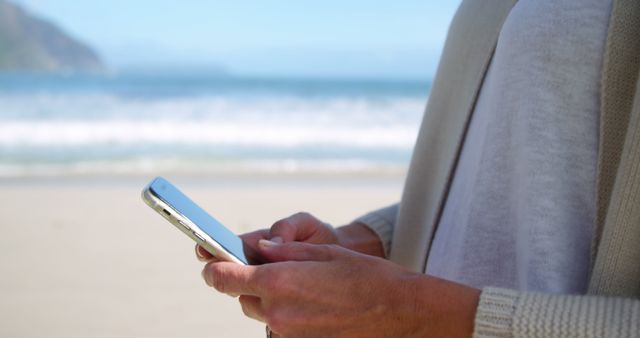 Person using smartphone while standing on beach on sunny day. Ocean waves in background, suggesting a moment of relaxation and connection to nature while staying connected to technology. Ideal for designs that require visuals related to modern lifestyle, remote work, or recreational activities.