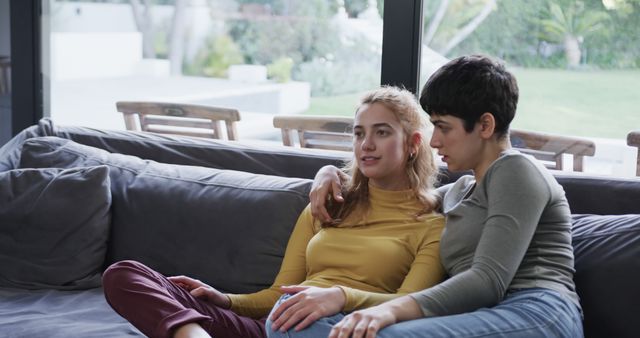 Two young women are sitting together on a modern sofa in a relaxed manner, with one woman embracing the other. They seem to be enjoying each other's company in a contemporary, well-lit home with a view of a garden through large windows. This visually appealing scene can be used for depicting themes of friendship, companionship, relaxation, modern living, and interaction at home. It is suitable for lifestyle blogs, interior design magazines, and social media posts focusing on relaxation and home environments.