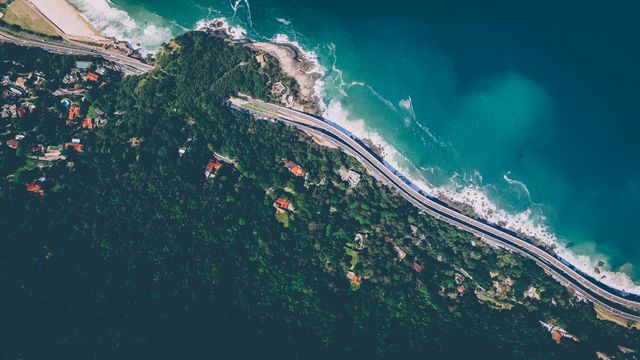 Dynamic overhead view highlighting the contrast between winding coastal road and lush green forested cliff by the blue ocean. Perfect for travel blogs, nature photography portfolios, and tourism advertisements. Useful for discussions on road trips, exploring nature, and coastal adventures.