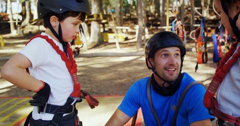 two individuals are in an adventure park setting, wearing safety equipment and engaging in what seems to be a preparatory discussion before starting an exciting outdoor activity. the scene captures the mix of fun and safety synonymous with nature adventure sports. this visual is ideal for promoting outdoor adventure clubs, safety gear for activities, or family-friendly adventure experiences.
