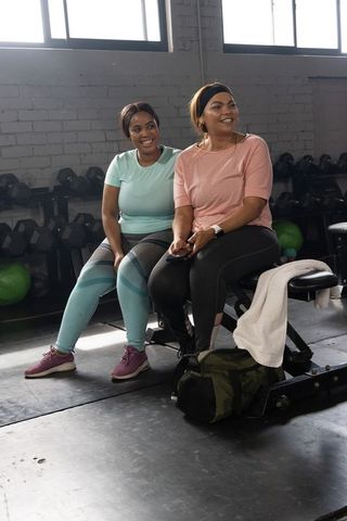African american women sharing workout break on bench in sunlit gym with smartphone