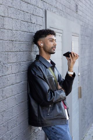 Asian man leaning on gray brick wall speaking into smartphone wearing leather jacket