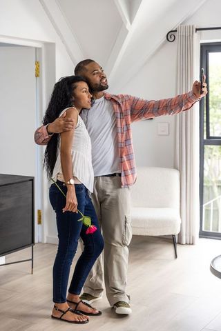 Diverse couple taking selfie in bright modern living room holding rose and embracing