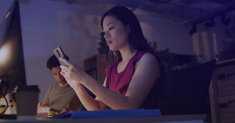 Mid adult woman working late scrolling smartphone in dim coworking office with monitor