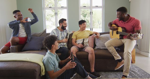 Group of young adults hanging out at home, relaxing and enjoying each other's company. One person is playing guitar while others have drinks in hand, smiling and engaging in light conversation. Great for concepts of leisure time, friendship, music sessions, and multicultural gatherings.