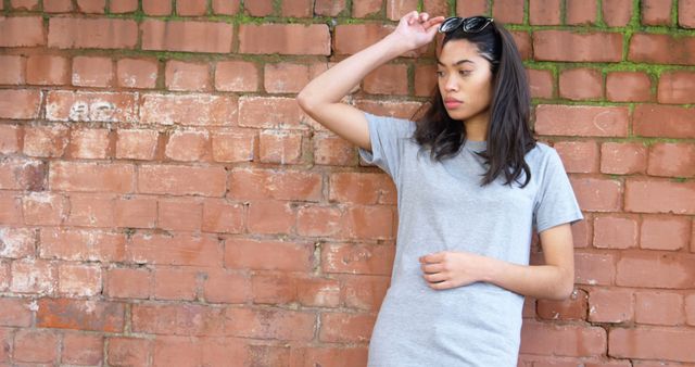 Young woman wearing gray t-shirt standing against rustic brick wall in casual pose. She holds sunglasses with one hand, looking relaxed and thoughtful. Ideal for use in fashion blogs, casual wear advertisements, urban style campaigns, or social media content focusing on youth fashion and lifestyle.