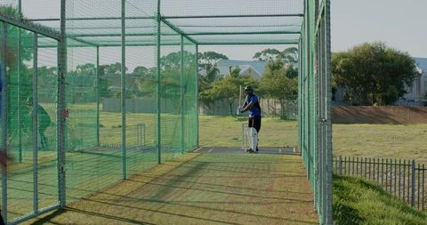 Cricket batsman practicing in outdoor net cage wearing helmet and pads on sunny field