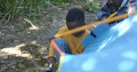 African american caregiver and boy securing blue tent at mossy campsite learning teamwork