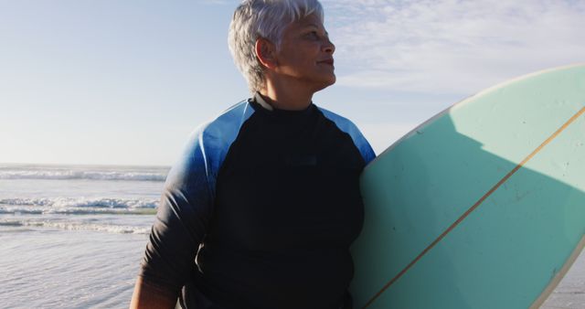 Senior woman holding a light blue surfboard on a beach with waves in the background. She wears a wetsuit and gazes into the distance with a calm expression in afternoon sunlight. Perfect for promoting active senior lifestyles, fitness and sport activities, seaside vacations, and personal growth over age.