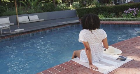 African american woman relaxing by backyard pool in white cover-up with straw hat and book