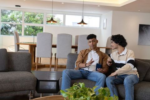 Two young men relaxing on sectional sofa using smartphone in sunlit modern living room