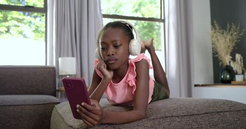Young girl listening with headphones while using smartphone on sunlit living room ottoman
