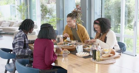 Diverse friends laughing and sharing homemade meal at sunlit wooden dining table