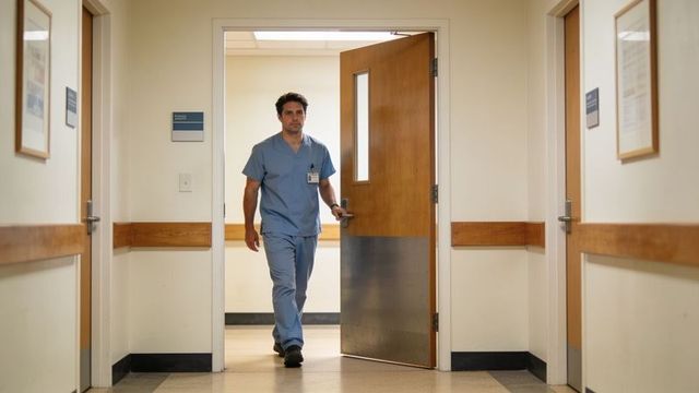 Walking clinician in blue scrubs entering hospital corridor with id badge