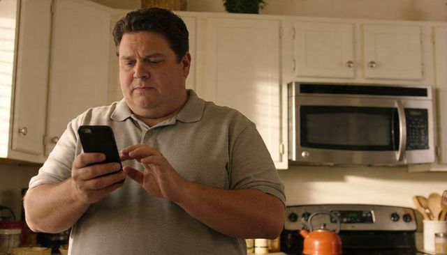Middle-Aged Man Using Smartphone in Bright Kitchen, Tapping Screen While Preparing Meal