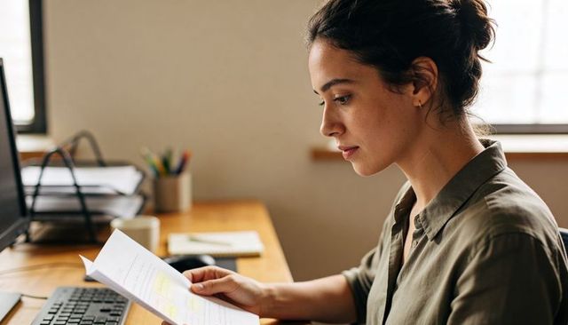 Focused young woman reading printed documents at home office desk in natural light