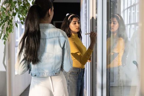 Multicultural women collaborating and writing on glass wall during brainstorming session