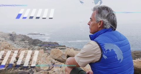 Senior man sitting on rocky seaside coastline wearing blue vest with digital hud overlays