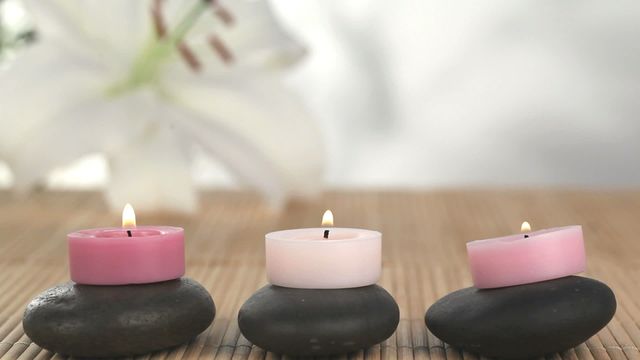 Three lit tea candles placed on smooth stones create a serene and calming ambiance shown against soft bamboo mat with elegant unfocused lily flower in the background. Perfect for brochures, websites, and advertising materials related to spas, wellness centers, and meditation practices, this image elegantly captures the essence of tranquility and peacefulness, making it ideal for promoting services or products aimed at stress relief and holistic well-being.