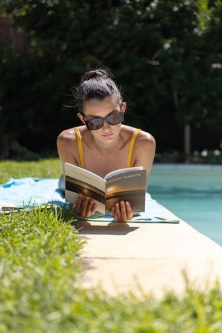 Woman reading endless sunsets while sunbathing on backyard pool deck wearing sunglasses