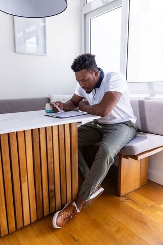 African american man working in sunlit kitchen nook checking planner and smartphone