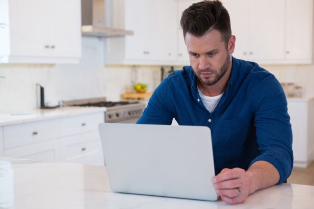 Man in casual blue shirt using laptop in modern kitchen. Ideal for content related to remote work, home office setups, technology use, and lifestyle blogs.