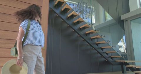 Woman turning in modern foyer with floating staircase, glass railing and straw hat