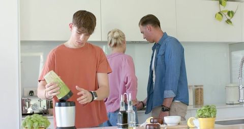 Teen making green smoothie at kitchen island while parents chat in bright modern kitchen