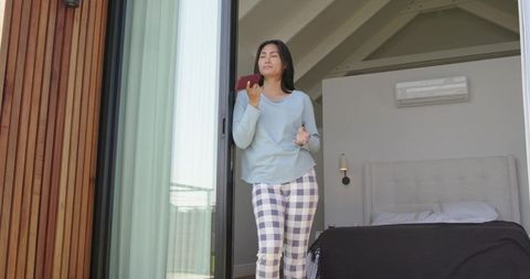 Asian woman standing in doorway wearing pajamas holding phone enjoying balcony morning
