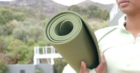 Indian woman carrying rolled green yoga mat outdoors on hillside balcony for wellness