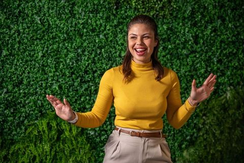 Smiling woman gesturing confidently, welcoming in mustard turtleneck before lush green foliage wall