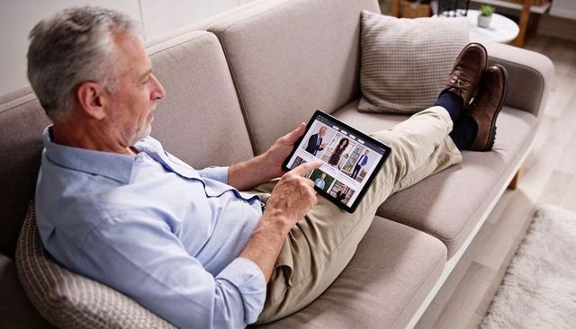 Senior man reclining on beige sofa using tablet for browsing, shopping, video calls