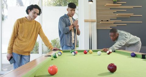 Diverse young men playing pool around green-felt table in sunlit game room