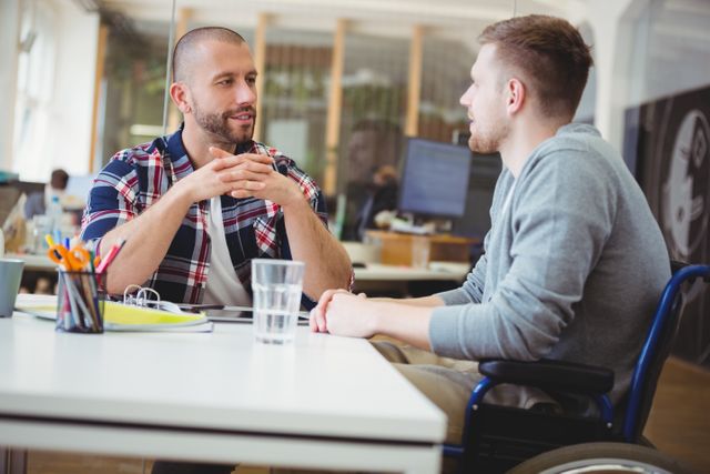 Young handicap businessman in wheelchair engaging in discussion with colleague in a modern, creative office. Ideal for illustrating concepts of teamwork, diversity, inclusion, and professional collaboration in a business setting.