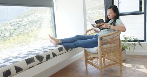 Asian woman relaxing in rattan chair reading paperback with feet on geometric window bench