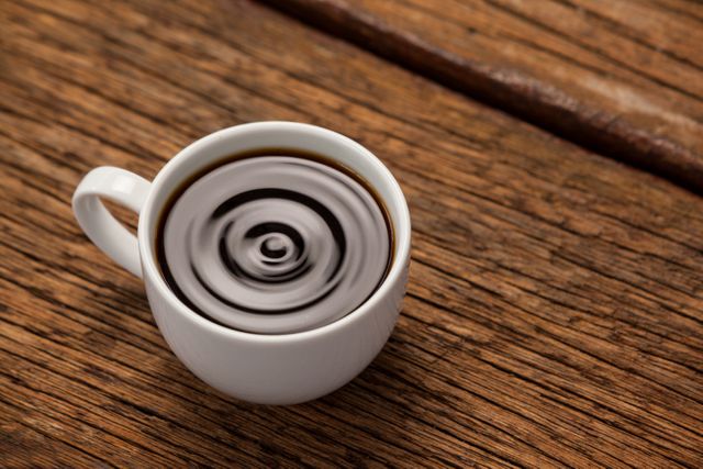 Close-up of a white coffee cup filled with black coffee, placed on a rustic wooden table. The coffee surface shows a ripple effect, adding a dynamic element to the scene. Ideal for use in articles about coffee culture, morning routines, relaxation, or rustic aesthetics. Suitable for blogs, social media posts, and advertisements related to coffee shops or cafes.