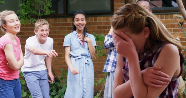 A group of diverse teenagers is laughing and pointing at another teenage girl who appears upset, covering her face with her hands. The scene captures a moment of peer pressure or bullying among young people, highlighting an emotional and challenging situation.