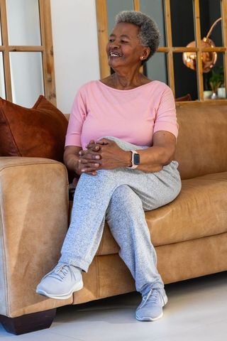 African senior woman relaxing on tan leather sofa wearing smartwatch in cozy living room