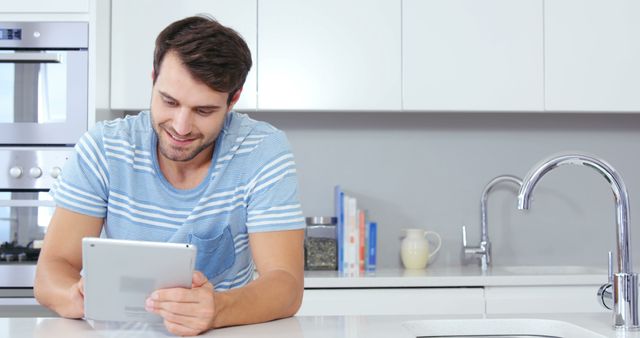 A man is using a tablet in a modern kitchen, engaging in work or leisure activities. The bright, contemporary kitchen features a sleek design and a clean atmosphere, emphasizing a comfortable and connected lifestyle. Ideal for use in content related to technology, home lifestyle, remote work, or modern home interiors.