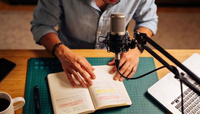 Recording podcast host speaking into condenser microphone with open notebook and laptop